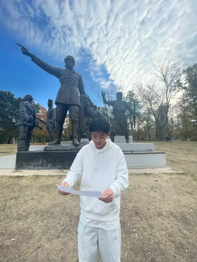 AIC student reading a printed sheet in front of the Kossuth monument in Orczy Park.